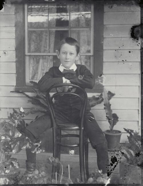 [Portrait of Ernest Withycombe astride a chair on verandah of timbered cottage with garden and pot plants] [picture] / Robert Morse Withycombe