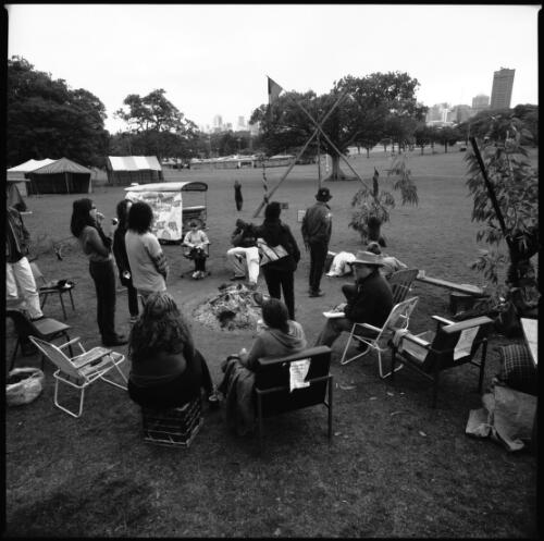 People sitting and standing around the sacred fire, the skyscrapers of Sydney CBD are in the background, Aboriginal Tent Embassy, Victoria Park, University of Sydney, 19 August 2000 [picture] / Loui Seselja