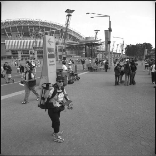 A woman carrying a large backpack in the shape of a Kodak roll of 35mm film sells film and other Kodak products to spectators. The Olympic Stadium is in the background, Sydney, 21 September 2000 [picture] / Loui Seselja