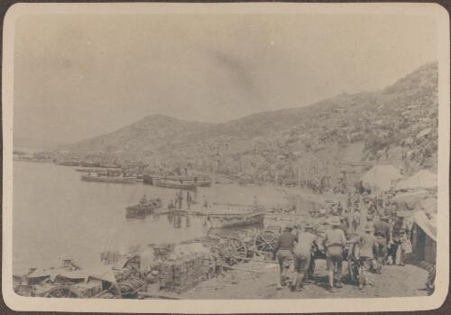 Beach scene, [at Anzac Beach where soldiers in the foreground push a loaded cart along a promontory lined with tents and fortifications and in the background boats are carefully moored around the beach, May, 1915] [picture] / J.P. Campbell