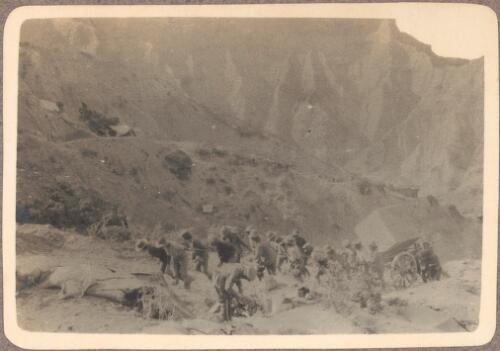 Further progress with tank [up the hillside to the headland above Anzac Cove, a line of about 16 soldiers drag a large cube-shaped tank on a two wheel cart with 3 or more pushing the cart from behind, against a rugged mountainous backdrop, May? 1915] [picture] / J.P. Campbell