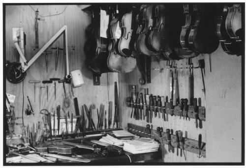 Numerous tools surround a work bench, while overhead hang violins awaiting repair, 1996 [picture] / Ian Kenins