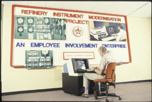 An unidentified Caltex employee sitting at a computer terminal of the digital control system in front of a display board showing examples of refinery instrument modernisation [transparency]