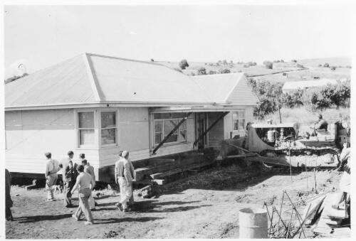 Workmen watch a Cat D8 bulldozer begin to push a house backwards on timber runners to make way for a Flagstaff service station at Darlington, South Australia, September 1956 [picture]
