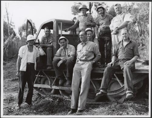 Nine men, including Project Engineer, Jack Cook, standing centre, and Bombardier driver, Stan Reynolds, seated in cabin, with a Bombardier vehicle, Hydro-Electric Commission expedition by Bombardier to the Serpentine River, Tasmania, 1961 [picture] / Russ Ashton