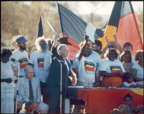 Professor Ovington and Mr. Reggie Uluru hold aloft copies of the lease aggreement to the applause of traditional owners; seated are Sir Ninian Stephen (partly obscured) and Mr. Holding [picture] / prepared by Media and Communications, Department of Aboriginal Affairs