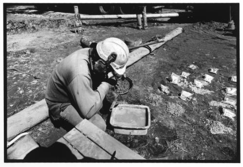 A hydrogeologist, tastes stones coming out of the bore, knowing from samples and the taste of minerals, how much deeper the bore has to be, 50 km from McCosker Bore Camp, 4 August, 2002 [picture] / Anya van Lit