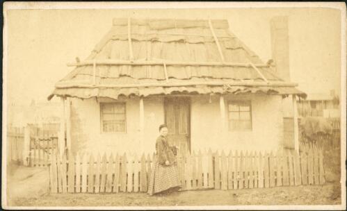 Pregnant woman at the front gate of a rendered cottage with bark roof and picket fence, Hill End, New South Wales, ca.1872 [1] [picture]
