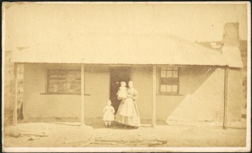 Woman with two young children under veranda of a rendered cottage with corrugated iron roof and goods for sale in left window, Hill End, New South Wales, ca.1872 [1] [picture]