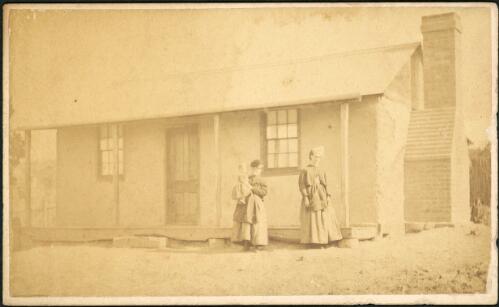 Two women and one child outside a rendered cottage with corrugated iron roof and brick chimney, Hill End, New South Wales, ca.1872 [1] [picture]