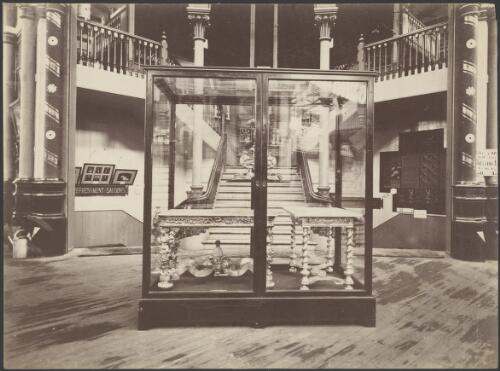 Ornate tables in a glass display case at the foot of a staircase in the Sydney International Exhibition, ca. 1880 [picture] / Richards & Co