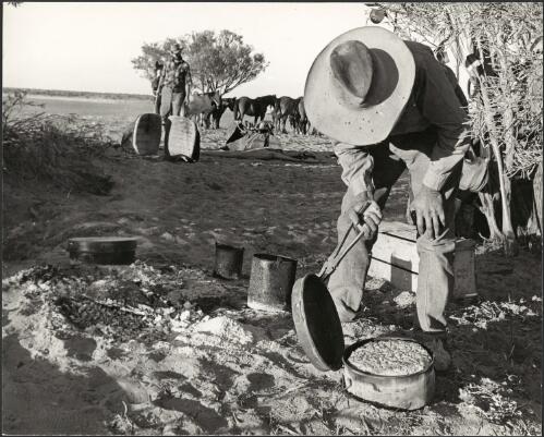 Drover and cook Azzi Fazulla uses pliers to lift the lid on the camp oven with the cooked damper,  Starvation Lake, South Australia, 1963 [picture] / Jeff Carter
