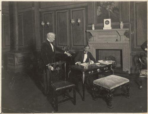 [Two male performers in formal dress with one standing and pointing at the other seated at a writing desk., in unidentified production between 1911 and 1925] [picture] / Sears' Studios, Melbourne