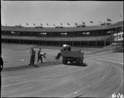 Groundsmen raking sand or gravel, which is being shovelled out of a truck onto the arena, Olympic Games, Melbourne, 6 December 1956 [picture]