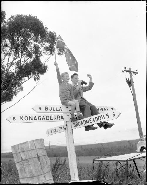 [Brothers John and David Gratton sitting on top of a road sign situated between Broadmeadows, Bulla, Konagaderra and Mickleham, to watch the 116 mile (185.6 kilometre) road race, Olympic Games, 7 December 1956] [picture]