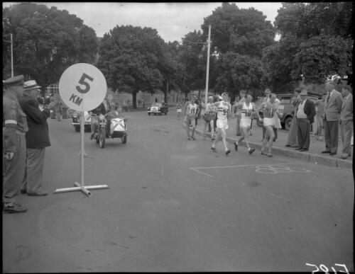[Competitors, including numbers 9, 10 G.W. Coleman (Great Britain), 11 Dordoni (Italy) and no.15 Hindmar (Sweden), being escorted by an official in a motorcycle sidecar, past the '5 km' sign in the 20 kilometres road walk, Olympic Games, Melbourne, 28 November 1956] [picture]