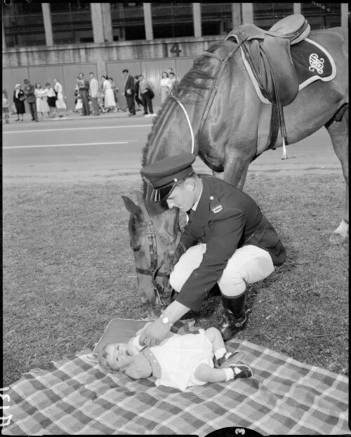 [Mounted policeman dismounted holding his horse's reins while bottlefeeding a baby on a picnic rug, Olympic Games, Melbourne, 22 November 1956] [picture]