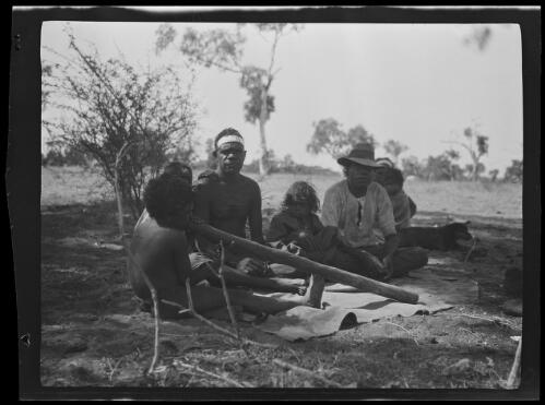 Two Aboriginal men, a woman, and children with a didgeridoo sitting on the ground, Wave Hill, Northern Territory, 1925 / Michael Terry