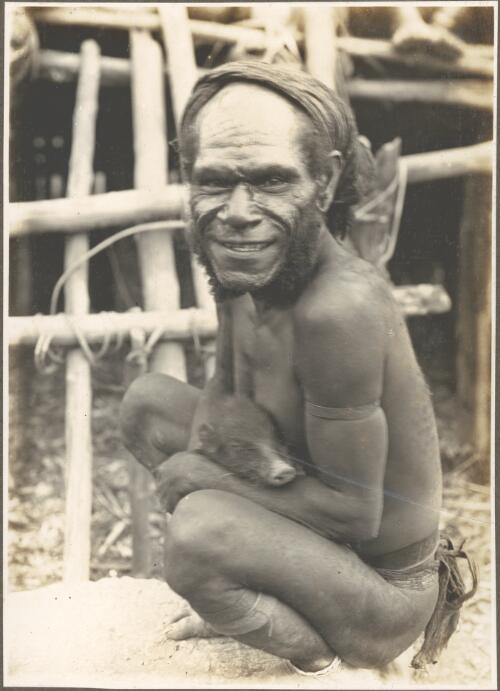 Types from villages of Evese, Ononge [man squatting and holding a pig] [picture]