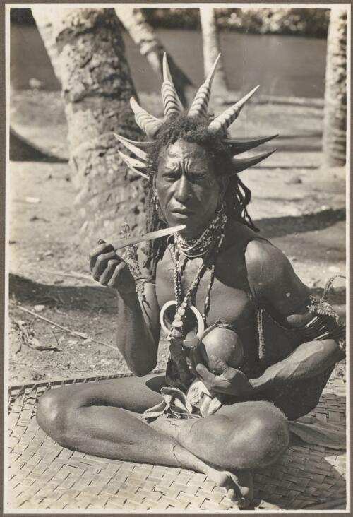 Wanigella [Wanigela] village [seated man wearing a head dress with large spikes, possibly eating betel nut] [picture] / Frank Hurley