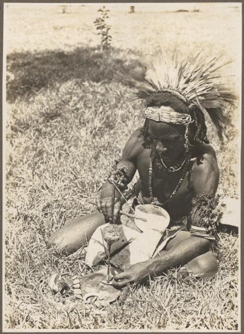 Buna Bay District [man seated on grass making a hole in a shell for a necklace] [picture] / Frank Hurley