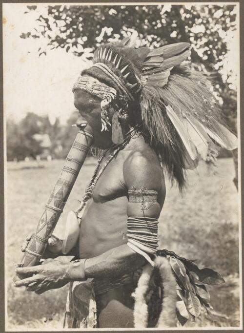 Buna Bay District [man in head dress with a carved wooden object at his mouth] [picture] / Frank Hurley