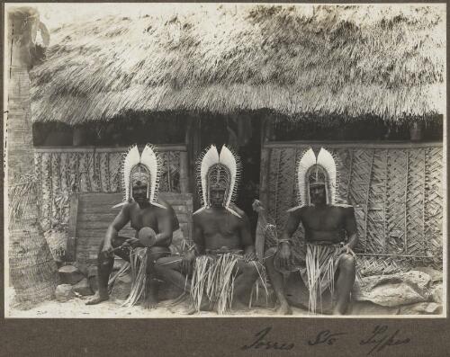 Torres Straits types [three men wearing head dresses sitting outside hut] [picture] / Frank Hurley