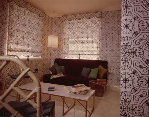 [Black and white wrought-iron-patterned paper is teamed with matching fabric blinds in this television room with cushions in emerald and peacock on a black textured ottoman with clear Perspex cube tables, ca. 1971] [transparency] / David Beal