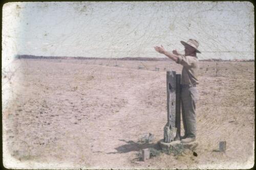 Man standing on a fence post at the corner of the border between South Australia and Queensland, known as Haddon Corner, inside the man's arms in South Australia, behind the man is Queensland, ca. 1950s [picture] / Les McKay