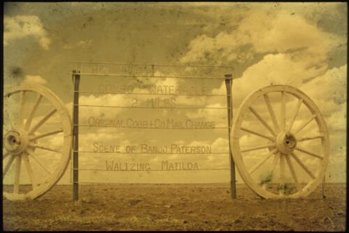 Sign flanked by wagon wheels, the sign reads, Winton Shire McKinlay Shire. Combo Waterhole 2 miles. Original Cobb + Co. Mail Change. Scene of Banjo Paterson Waltzing Matilda, Queensland, 1960s [transparency] / Les McKay