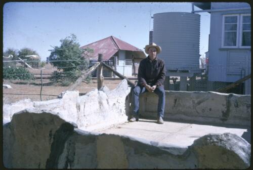 Man wearing a hat sitting on the edge of stone enclosure with a water tank and two buildings behind him [transparency] / Les McKay