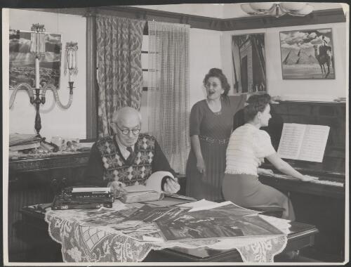 Frank Hurley reading a book with his daughters playing the piano and singing, 1950 [picture]