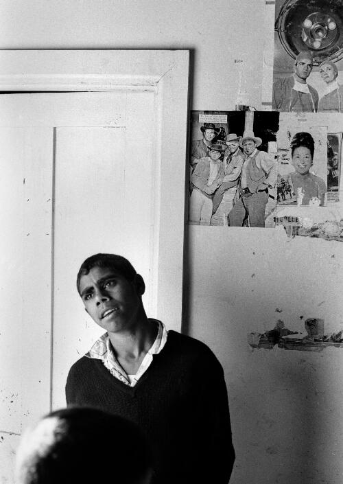 Young Aboriginal man standing in front of wall covered with posters of actors, Lake Tyers Mission, Victoria, ca. 1965 [picture] / Albert W. Brown