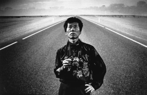 Photographer William Yang holding a camera with an image of a road being projected behind him, 1990s? [picture] / Robert McFarlane