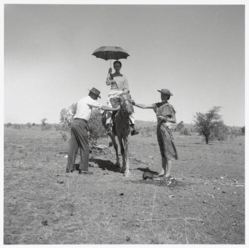 Cynthia Nolan mounted on the saddled dry carcass of a horse, with a man and a woman standing either side, Queensland, 1952 [picture] / Sidney Nolan