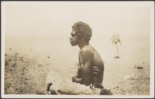 Young Aboriginal man sitting with his legs crossed and arms folded, with a camera on a tripod in the background, Gulf of Carpentaria, 1914 [picture] / Frank Hurley