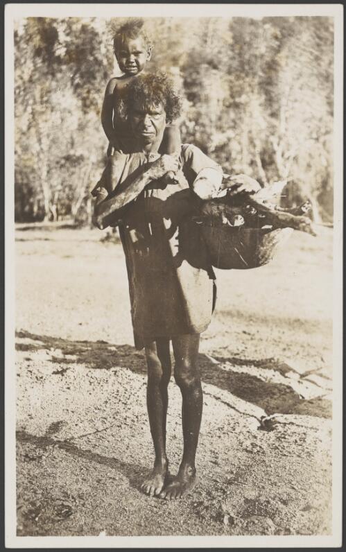 Aboriginal woman with a child sitting on her shoulders holding a woven bag of fire wood, 1914 [picture] / Frank Hurley
