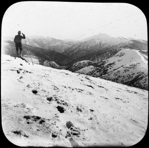 A trail of footsteps in the snow leading to a man with a pole looking at the mountain ranges, Victoria, approimately 1920 / Robert Wilkinson