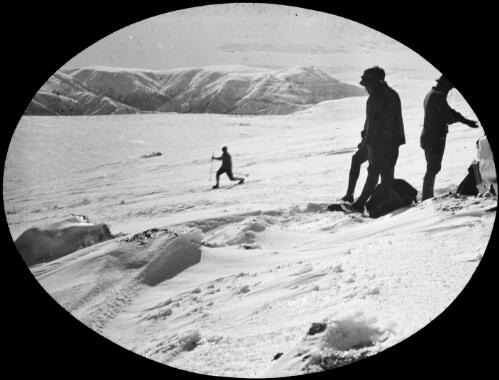 A man telemark skiing past a group of three men, Victoria, approximately 1920 / Robert Wilkinson