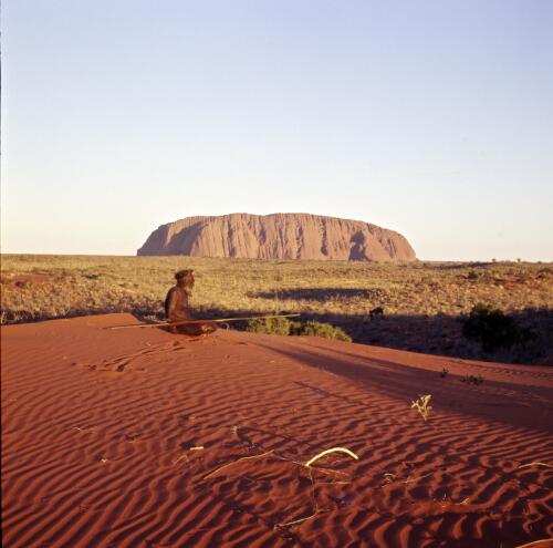 A Pitjantjatjara man sitting on a sand hill with Uluru in the background, Northern Territory, approximately 1966 / Robin Smith