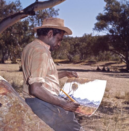 Aboriginal Australian stockman leaning against a rock and painting a landscape, Northern Territory?, approximately 1966 / Robin Smith