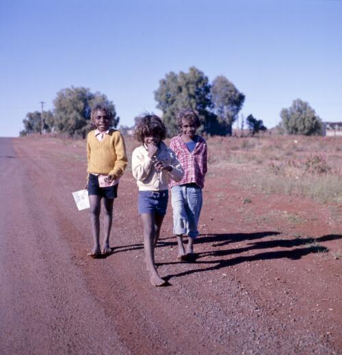 Three Aboriginal Australian school children walking along a dirt road, Leonora, Western Australia, approximately 1978 / Robin Smith