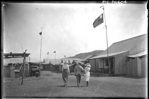 [Woman, a man and girl, all wearing hats, walking along a road of houses and flags, Darwin trip with Francis Birtles?] [picture] : [Darwin, Northern Territory] / [Frank Hurley]
