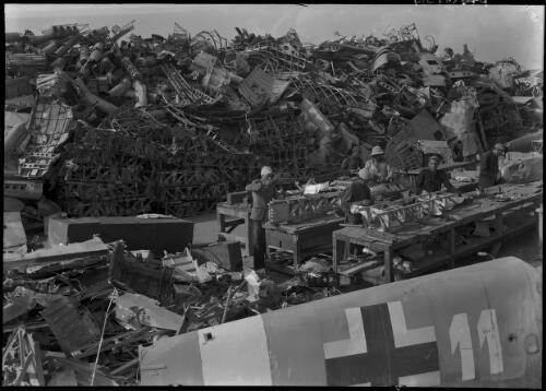 [Salvage dump, six figures at work benches, aircraft part with logo of cross and 11 and large pile of materials and equipment] [picture] / [Frank Hurley]