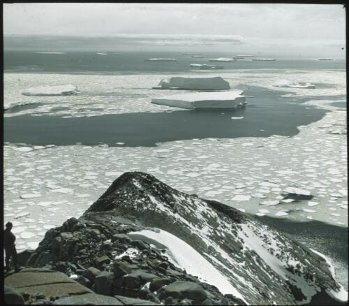 [Man standing on red rocks looking at view of water, ice and icefloes, Antarctica] [picture] : [Antarctica] / [Frank Hurley]