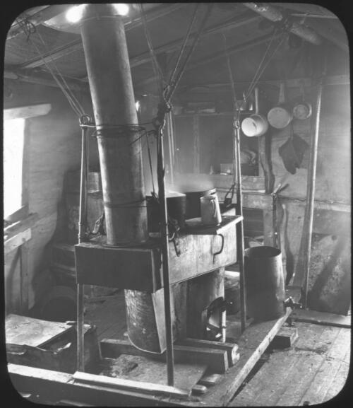 [A kitchen with a long stovepipe venting through the roof, saucepans, a window, a jug and mittens] [picture] : [Antarctica] / [Frank Hurley]