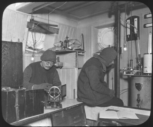 The Rookery, the scientific laboratory with Hussey [right] examining the Dimes anemometer and James [left] removing rime from the dip-circle, the electrograph is on the left [Shackleton expedition, 1914-1916] [picture] : [Antarctica] / [Frank Hurley]