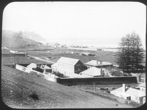 [A building surrounded by a high stone wall, two trees, a slope and the sea in the distance] [picture] : [Norfolk Island] / [Frank Hurley]