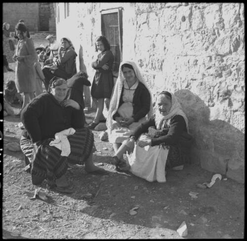 Jerusalem [women sitting on the ground and on benches near a stone wall and a girl wearing a short dress and stockings] [picture] / [Frank Hurley]