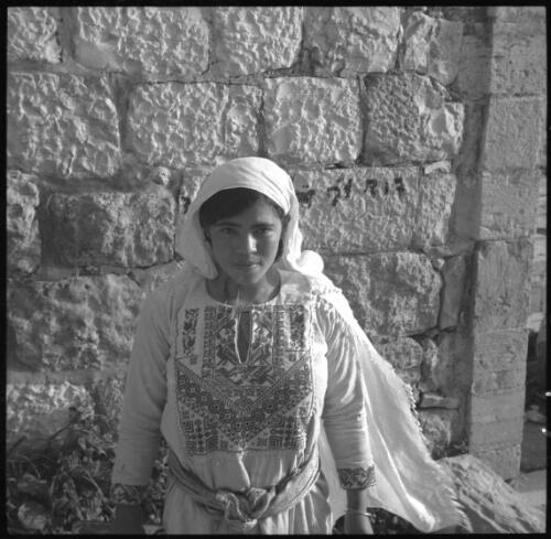 Few Jerusalem snaps [portrait shot of girl wearing patterned dress and fringed headshawl in front of a stone wall] [picture] / [Frank Hurley]